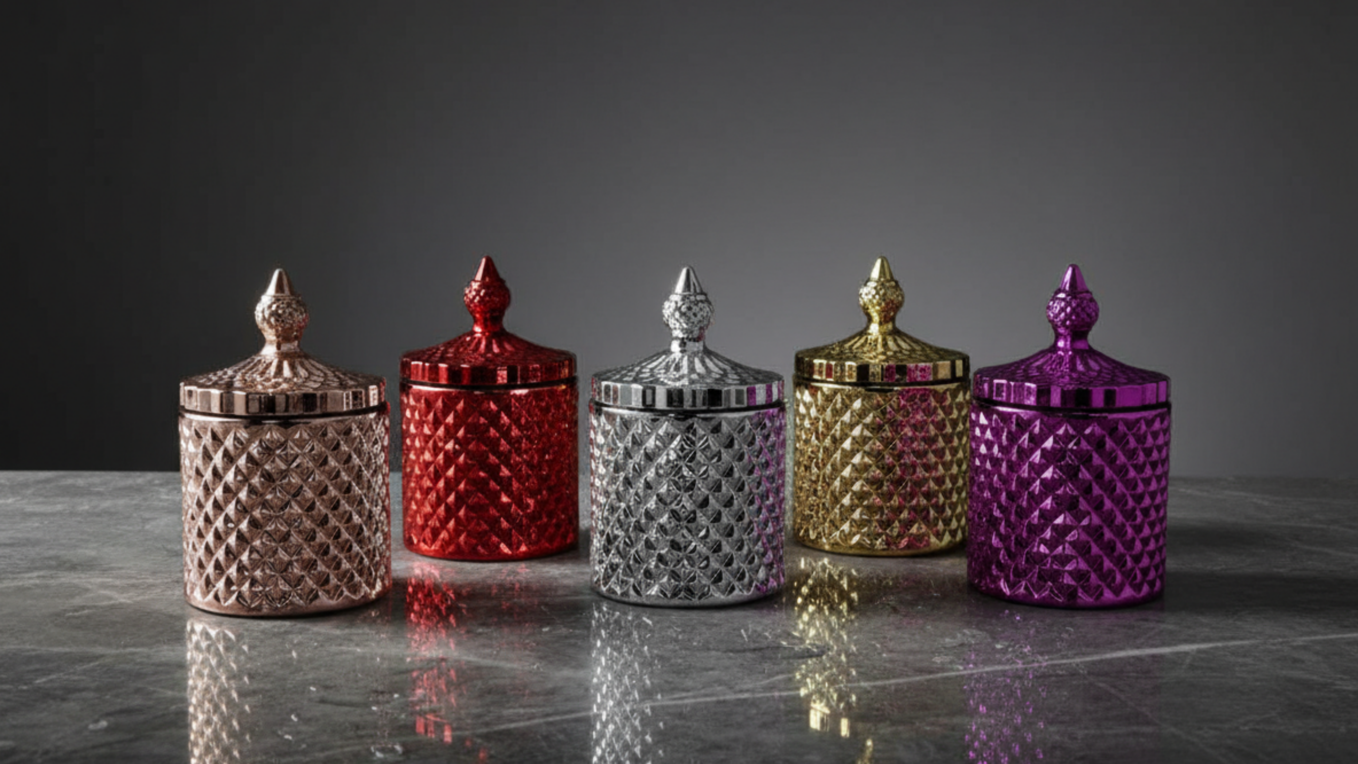 Five decorative jars with lids in rose gold, red, silver, gold, and purple on a reflective surface.