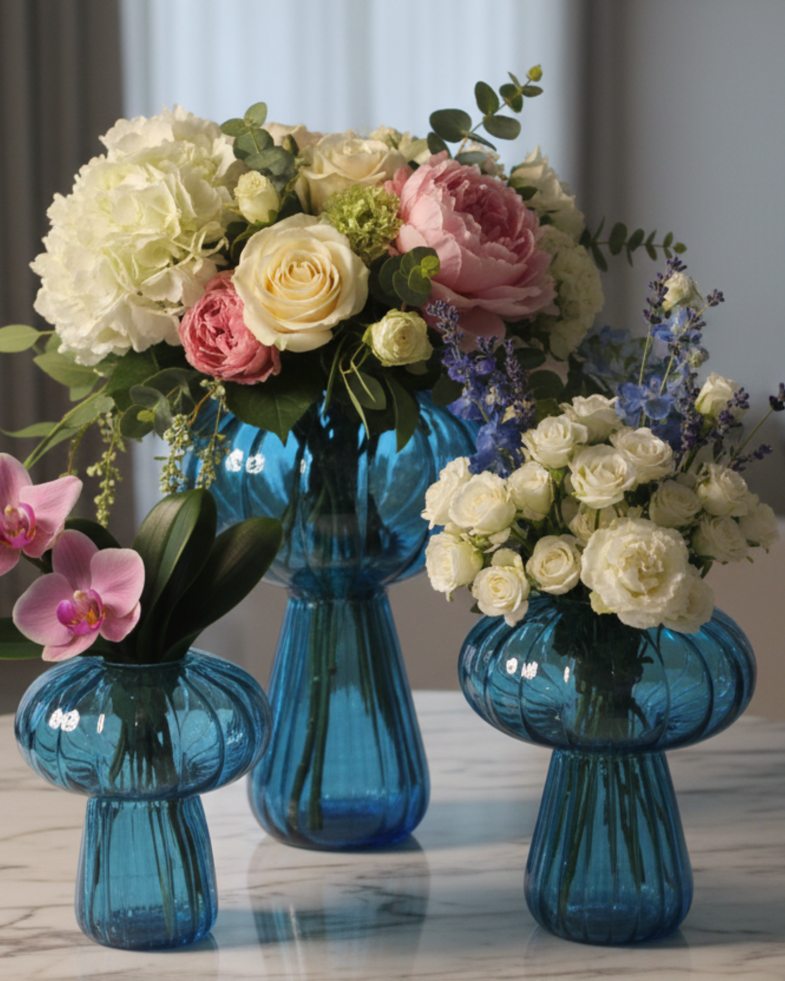 Three blue glass vases with floral arrangements on a marble surface.