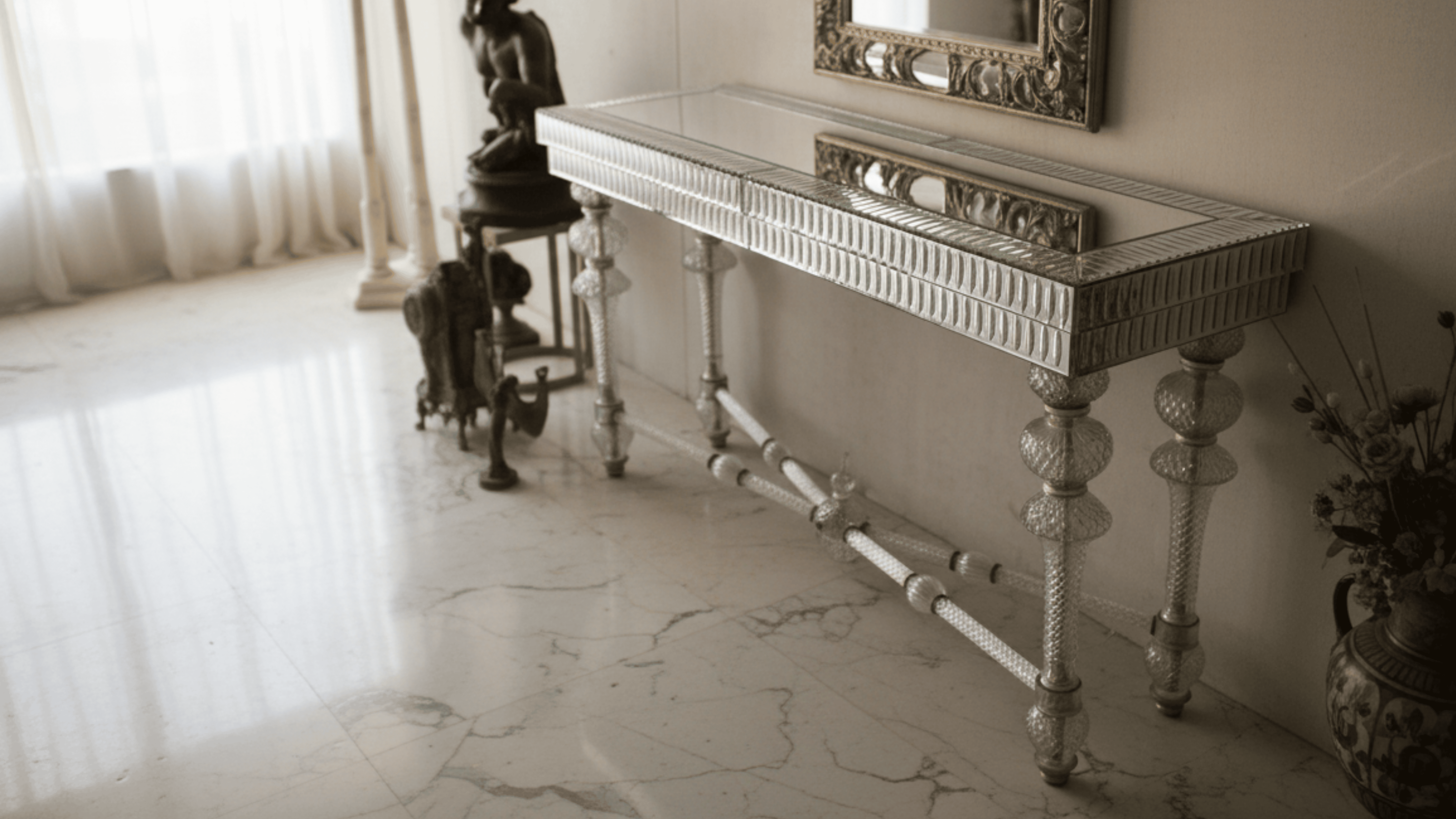 Decorative console table with intricate design in a room with marble floor and curtains.