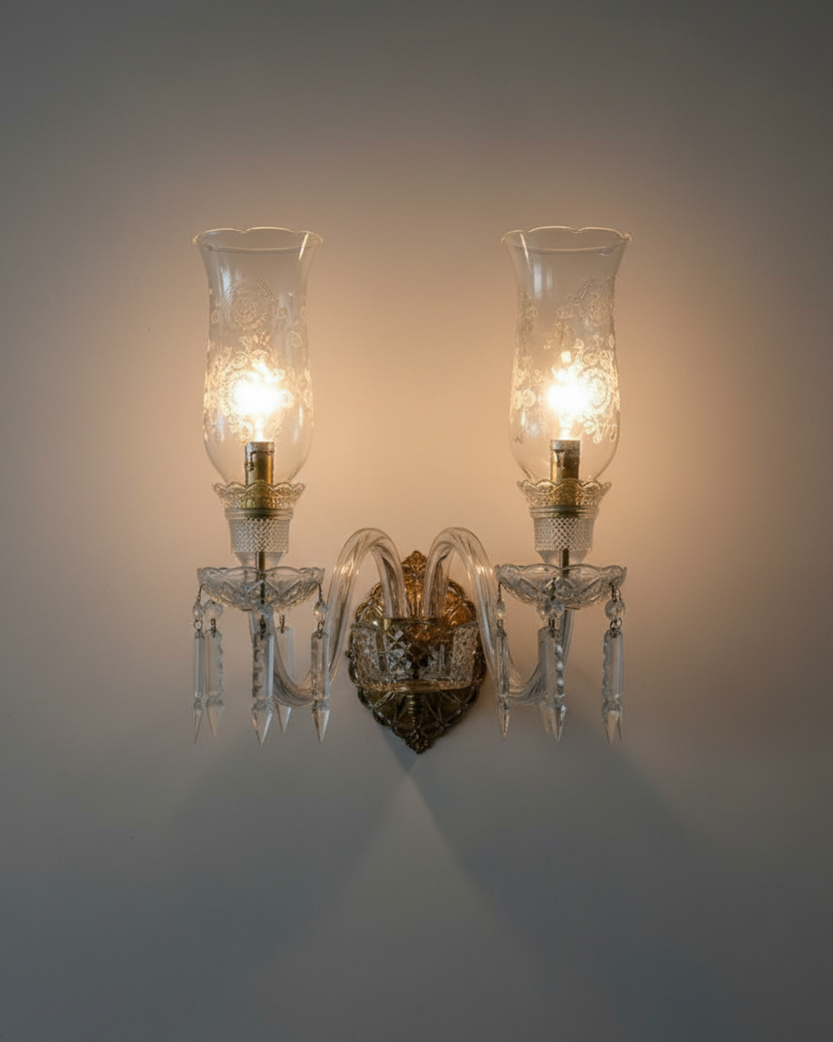 Pair of crystal wall sconces with glass shades on a plain wall. Handcrafted twin-arm wall light made in India.